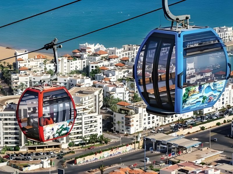 Two gondolas on a cable car system overlooking a coastal city with buildings and blue ocean in the background.