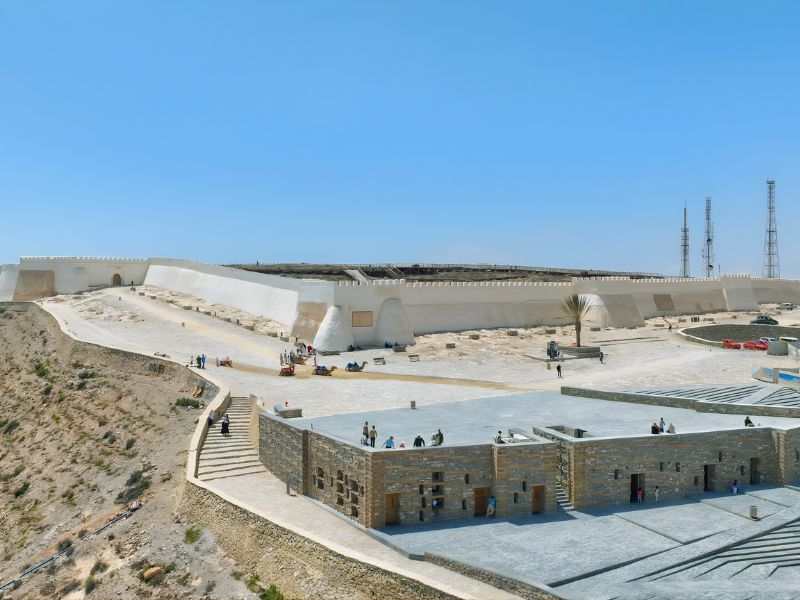 A panoramic view of a historical fort with white walls, set against a clear blue sky.Visitors are seen exploring the area, with modern structures visible in the foreground and communication towers in the background.