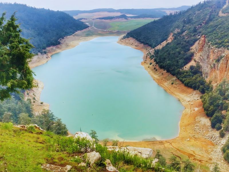 Aerial view of a serene lake surrounded by hills and greenery, with a sandy shoreline and distant fields in the background.