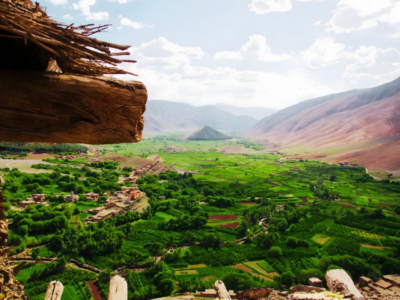 A panoramic view of a lush green valley surrounded by mountains under a partly cloudy sky, with a wooden structure in the foreground.
