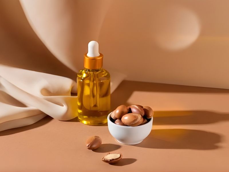 A glass dropper bottle filled with golden oil next to a small white bowl containing whole and broken argan nuts on a sandy surface.