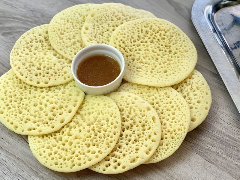 A plate of traditional Ethiopian injera, featuring multiple round, pale-yellow flatbreads with a spongy texture, arranged in a circular pattern with a small bowl of brown dipping sauce in the center.