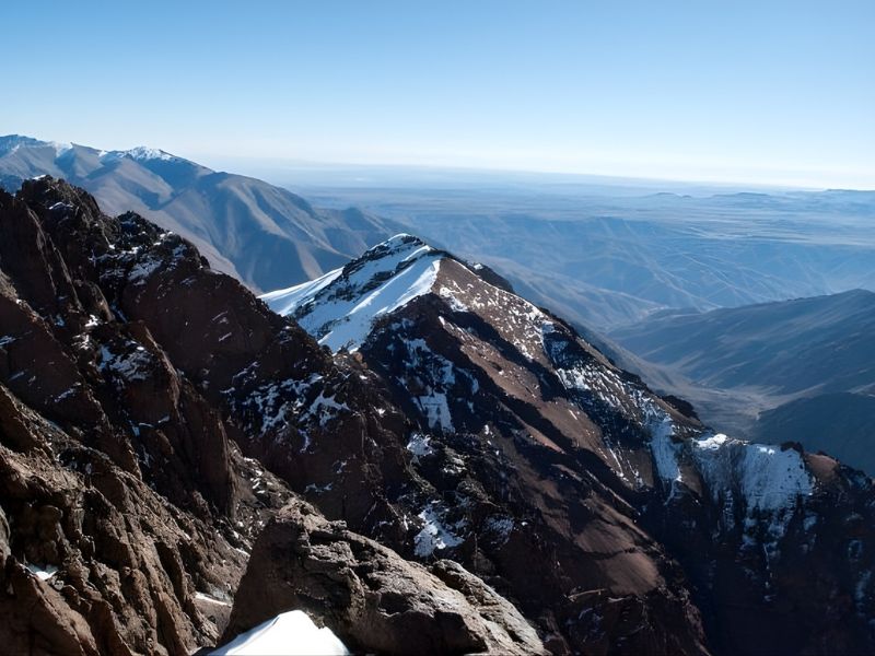 Panoramic view of snow-capped mountain peaks with rocky terrain and clear blue skies in the background.