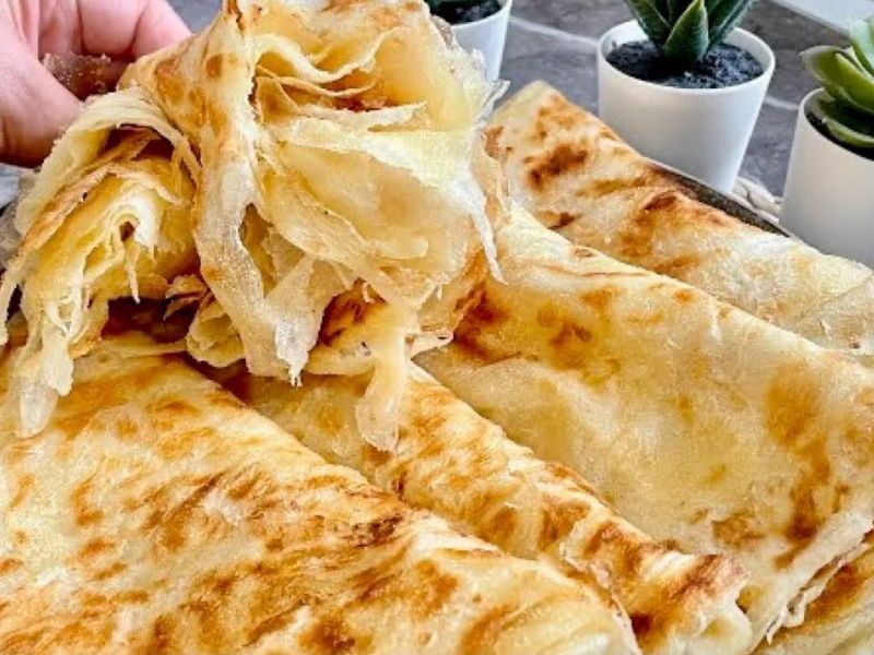 A close-up of freshly cooked, golden-brown flatbreads, with one piece being pulled apart to show its flaky texture. In the background, small potted plants add a decorative touch.