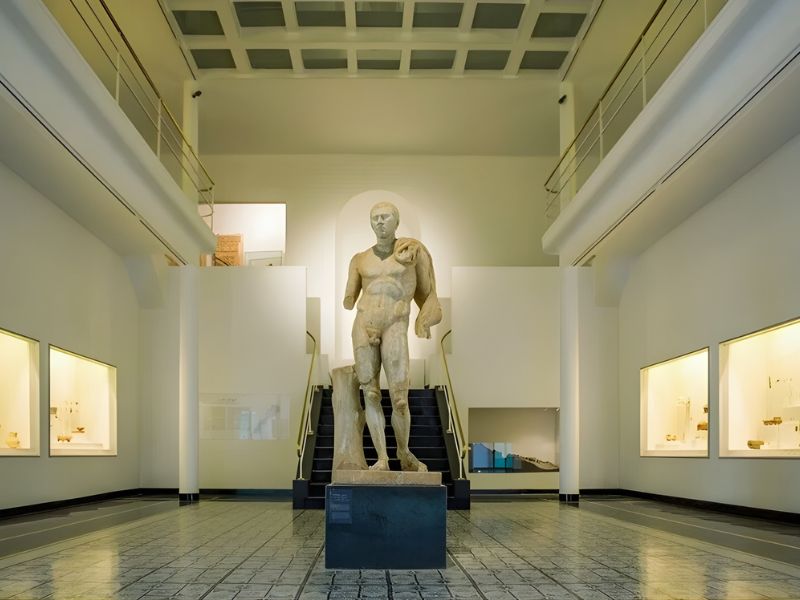 A large stone statue of a male figure stands prominently in a minimalist museum interior, featuring white walls, large windows, and well-lit display cases showcasing artifacts.