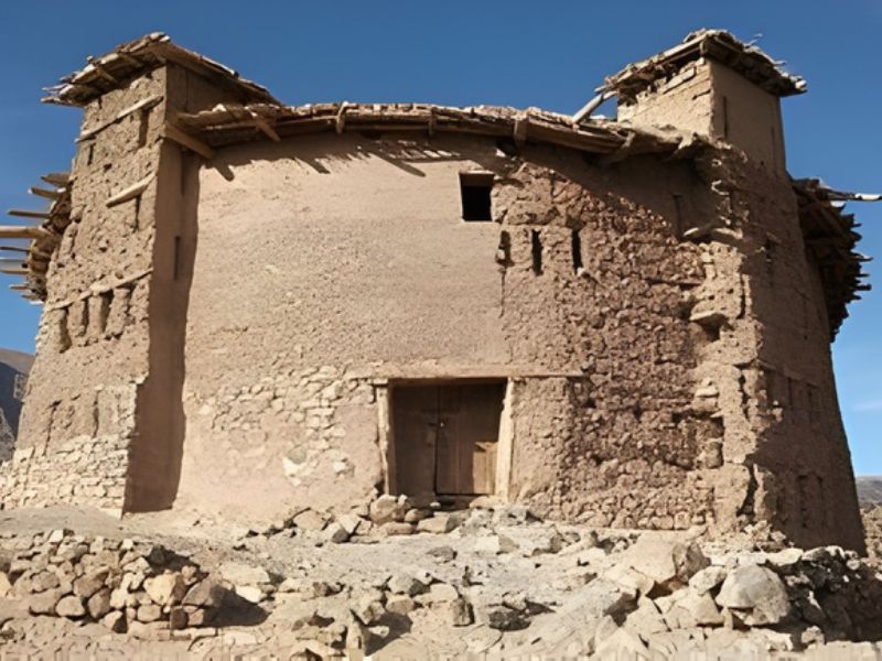 An ancient mud-brick structure with a flat roof and wooden beams, situated in a rocky environment under a clear blue sky.
