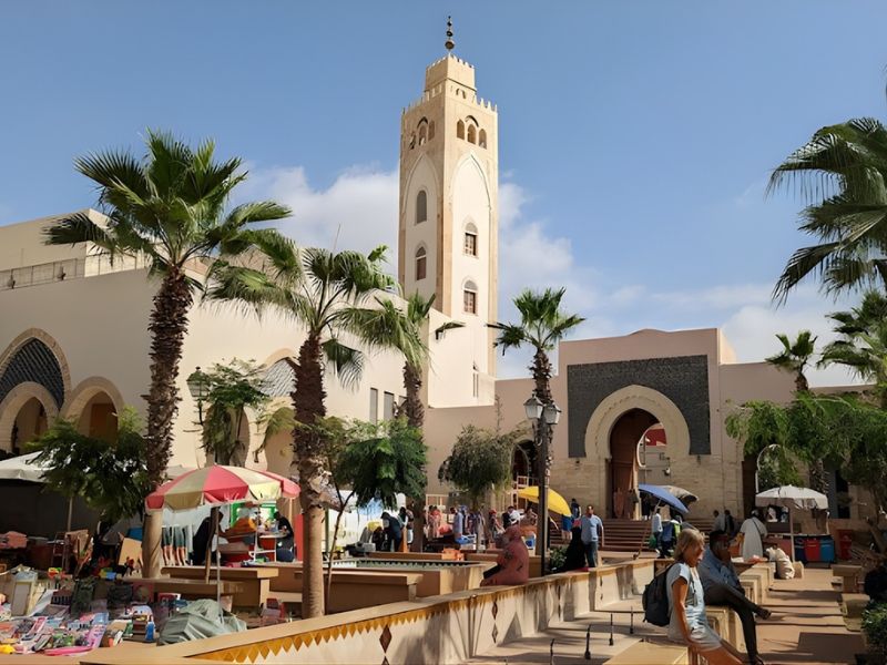 A vibrant marketplace featuring palm trees and colorful umbrellas, with a historic tower in the background under a clear blue sky.