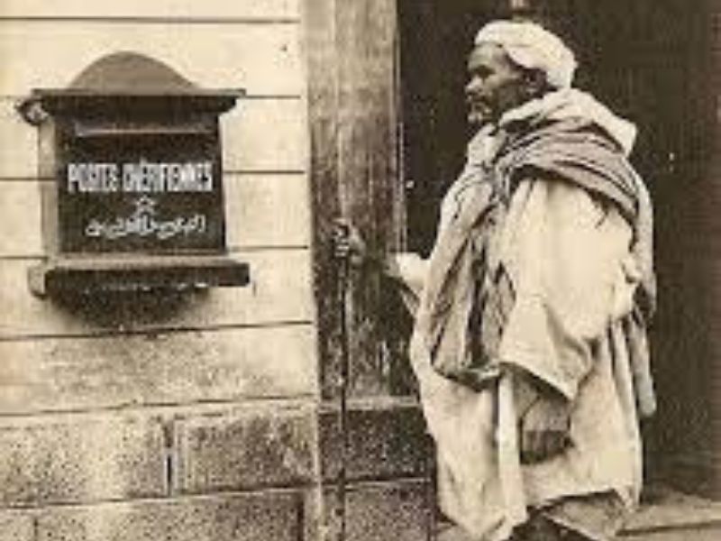 An elderly man in traditional attire stands next to a wooden door with a sign that reads 'Postes Cherifiennes' in French and Arabic.