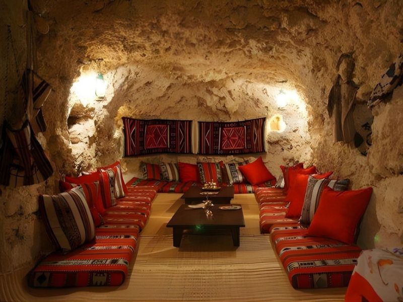 Interior of a cozy cave-like room with stone walls, featuring colorful patterned seating and cushions, a low wooden table, and decorative textiles on the walls.