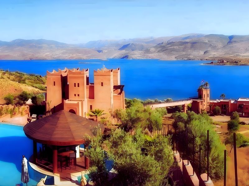 A historic adobe-style building near a calm blue lake with surrounding hills and greenery