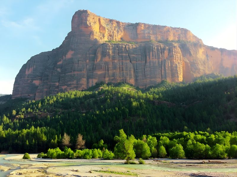 Large rock cliff with vertical striations towering above a dense green forest and a riverbed